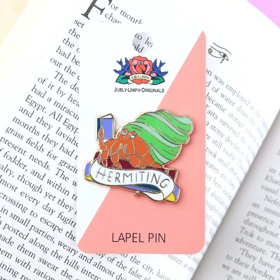 A hard enamel lapel pin on a Jubly-Umph backing card. The pin is in the shape of a Hermit Crab reading a book. The pin says Hermit Crab.