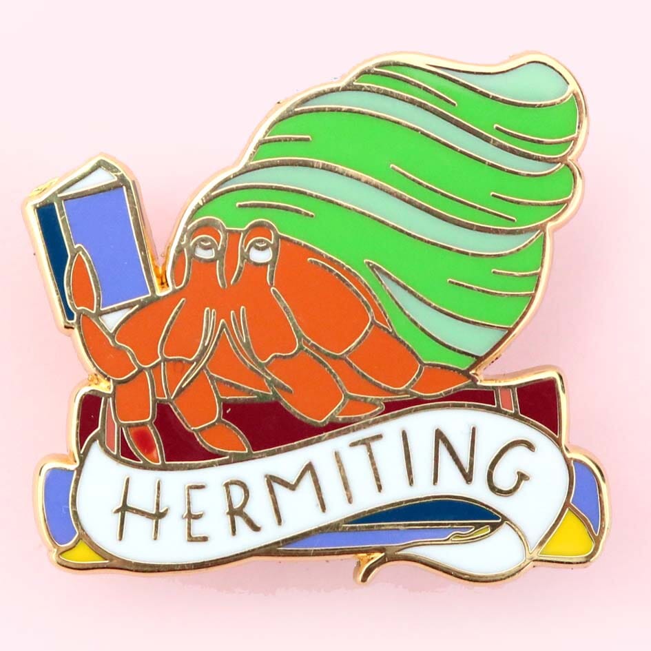 A hard enamel lapel pin on a pink background. The pin is in the shape of a Hermit Crab reading a book. The pin says Hermit Crab.