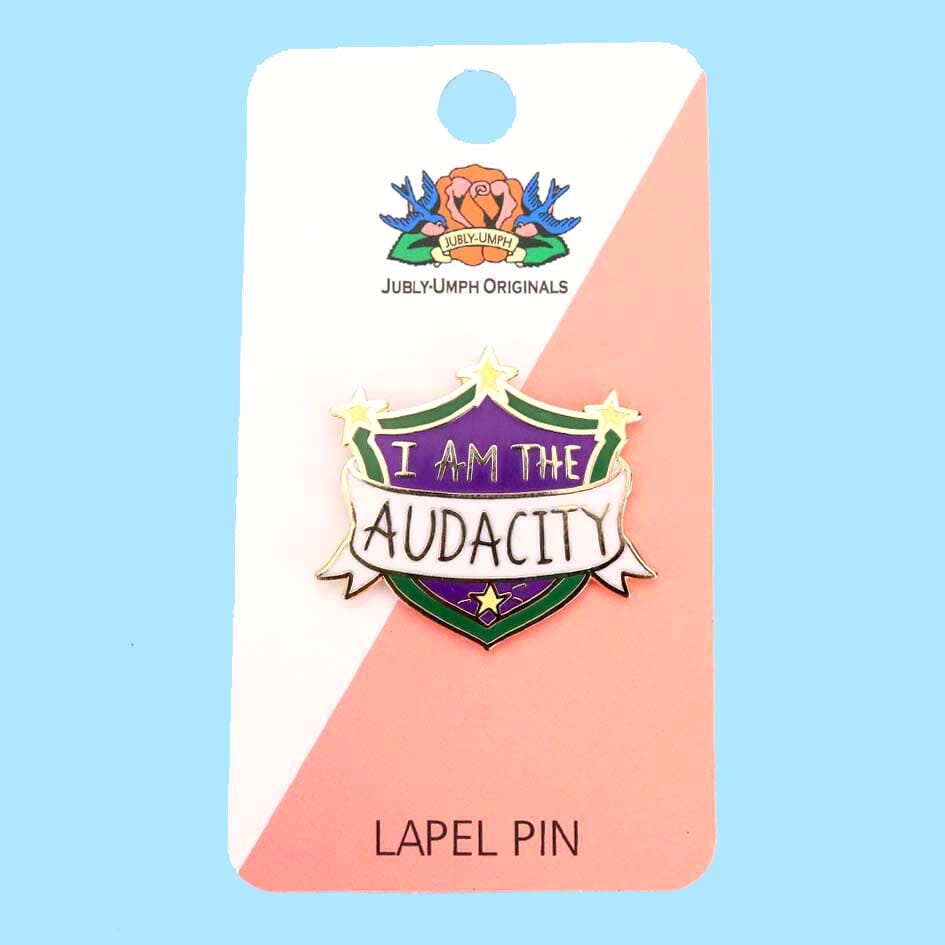 A hard enamel lapel pin on a Jubly-Umph backing card. The pin is in the shape of a shield. The lapel pin reads I am the Audacity.