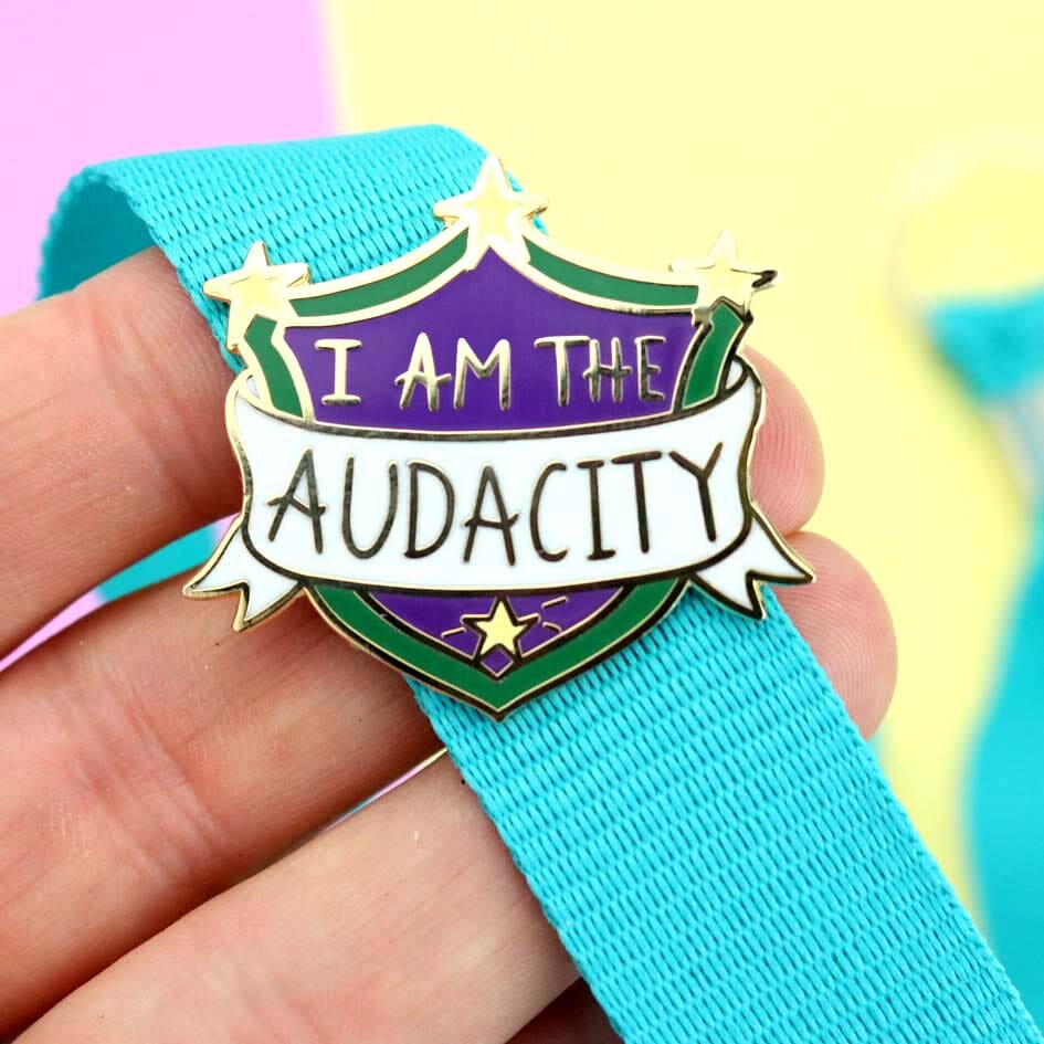 A hard enamel lapel pin being held in a hand on a blue lanyard. The pin is in the shape of a shield. The lapel pin reads I am the Audacity.