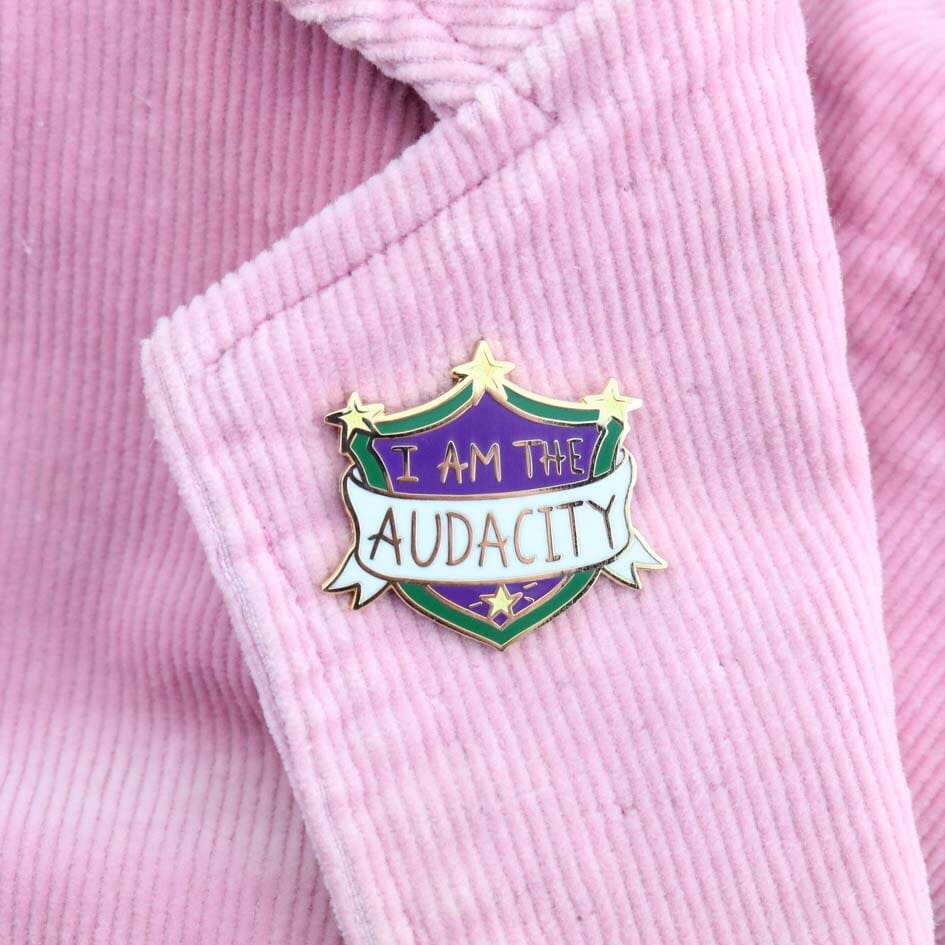 A hard enamel lapel pin being held in a hand on a pink Jacket. The pin is in the shape of a shield. The lapel pin reads I am the Audacity.