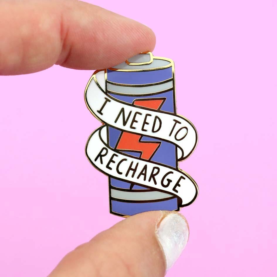 A hard enamel lapel pin held in a hand. The lapel pin is in the shape of a battery and reads I Need To Recharge.