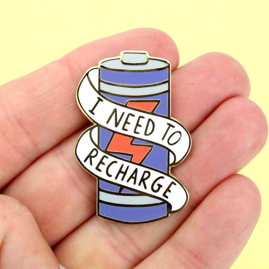 A hard enamel lapel pin held in a hand. The lapel pin is in the shape of a battery and reads I Need To Recharge.