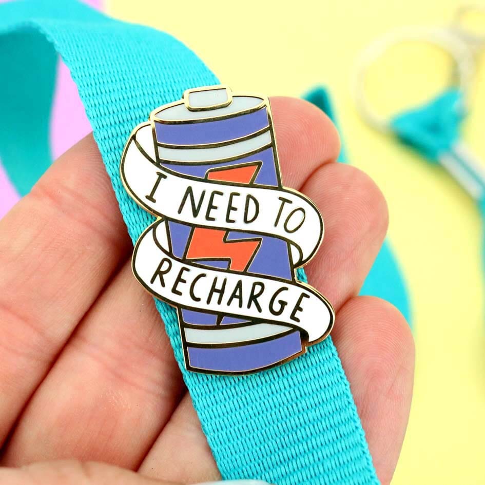 A hard enamel lapel pin being held in a hand on a blue lanyard. The lapel pin is in the shape of a battery and reads I Need To Recharge.