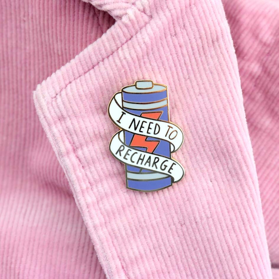 A hard enamel lapel pin on a pink jacket. The lapel pin is in the shape of a battery and reads I Need To Recharge.