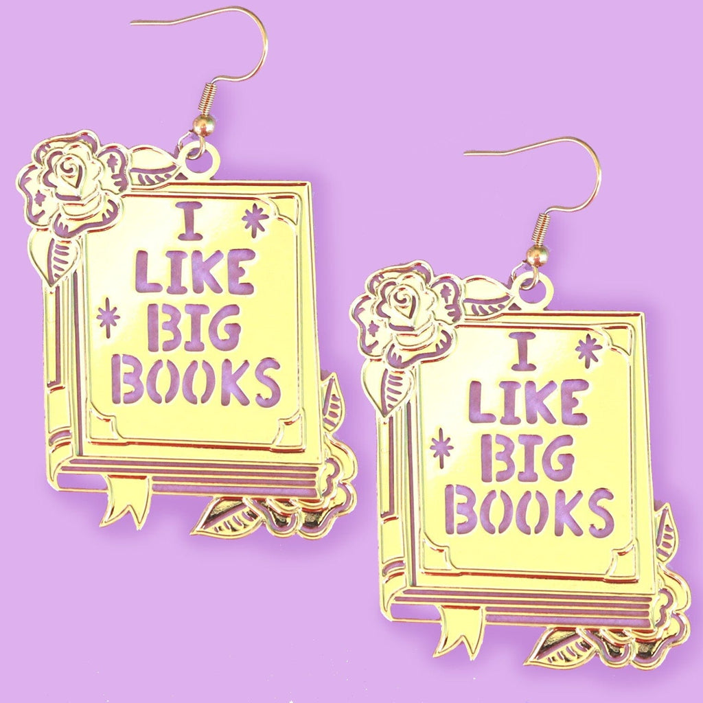 A pair of dangle brass earrings displayed on a purple background. The earrings read I Like Big Books.