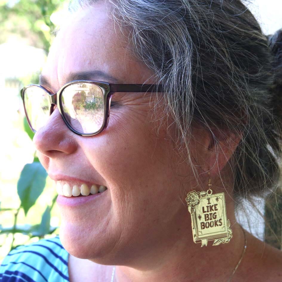 A single brass earring being worn by a model. The earring reads I Like Big Books.