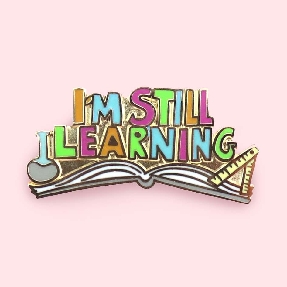 A hard enamel lapel pin against a pink background. The lapel pin is an open book and reads I'm Still Learning.