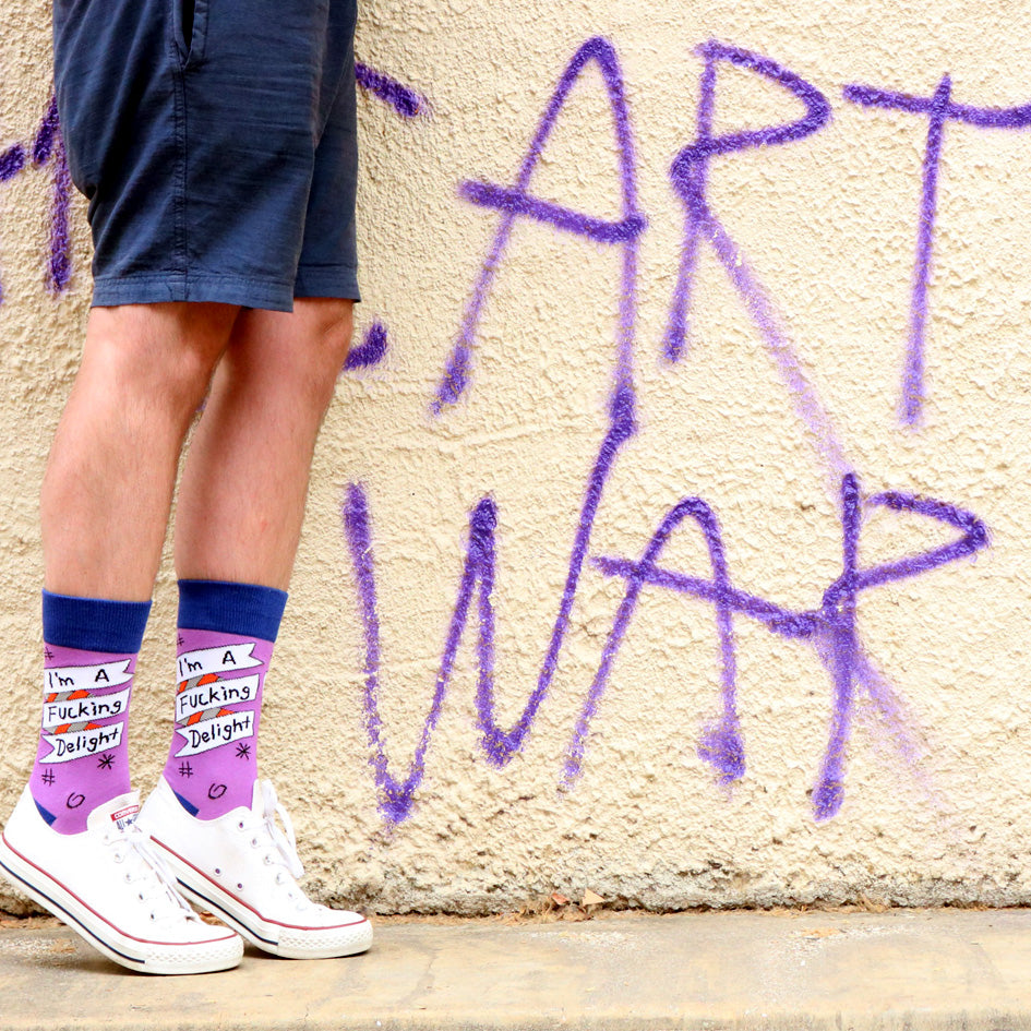 A pair of socks being worn with white shoes against a graffiti wall. The socks are purple and blue and read I'm A Fucking Delight.