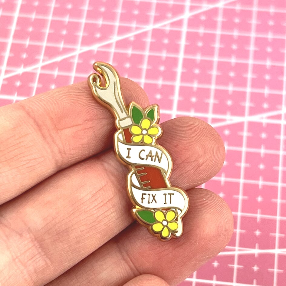 A hard enamel lapel pin being held in a hand. The pin is in the shape of a seam ripper. The lapel pin reads I Can Fix It.