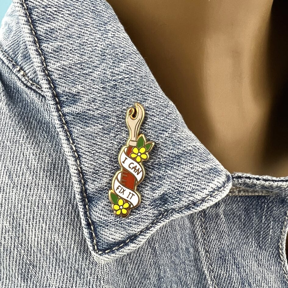 A hard enamel lapel pin being displayed on a denim jacket. The pin is in the shape of a seam ripper. The lapel pin reads I Can Fix It.