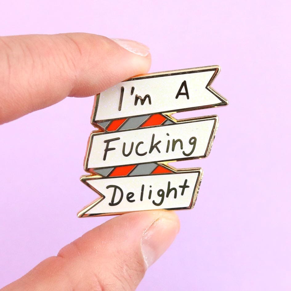 A hard enamel lapel pin being held in a hand. The lapel pin reads I'm A Fucking Delight.