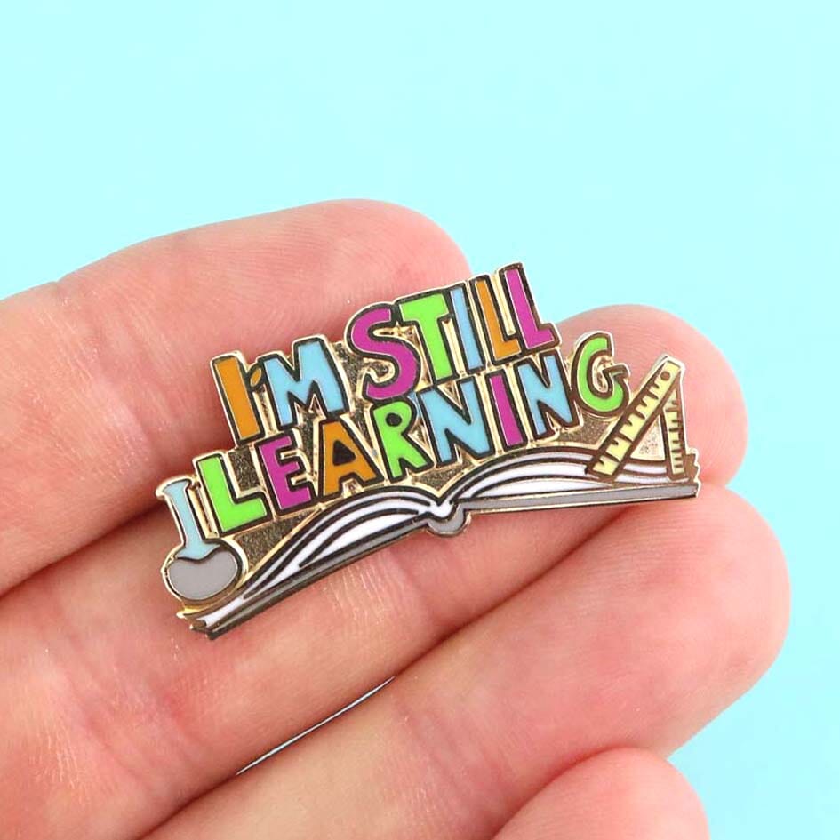 A hard enamel lapel pin being held in a hand. The lapel pin is an open book and reads I'm Still Learning.