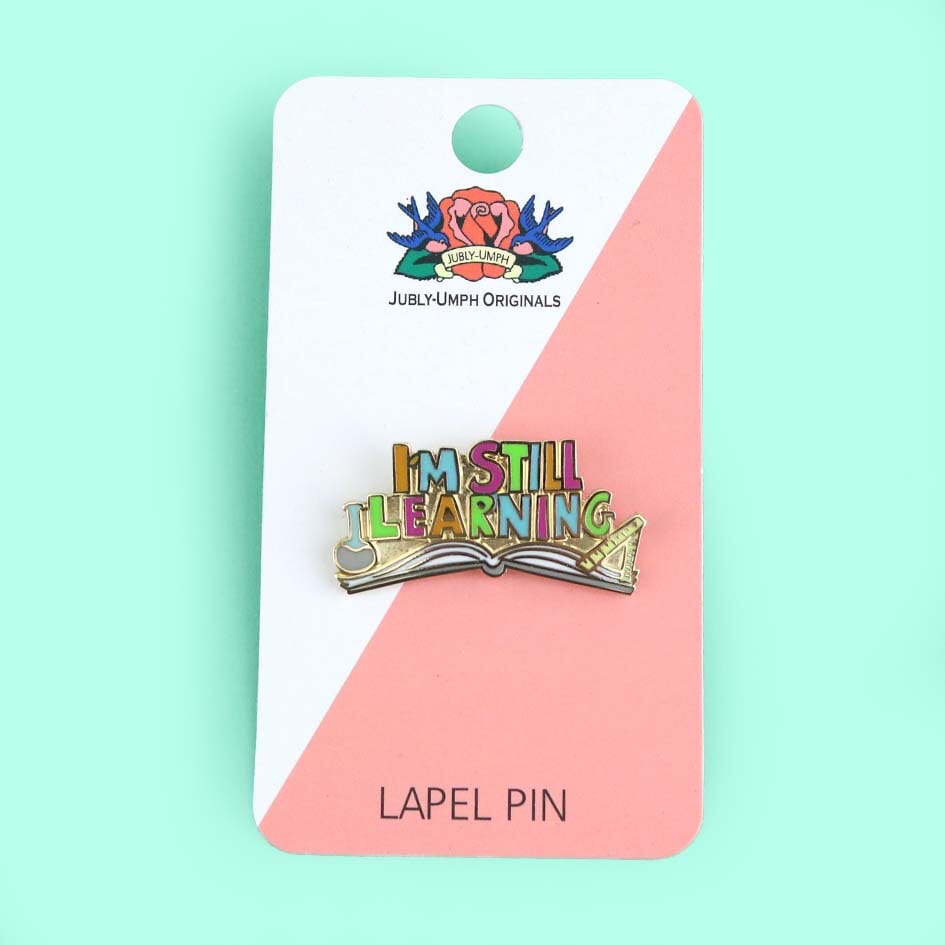 A hard enamel lapel pin being displayed on a Jubly-Umph backing card. The lapel pin is an open book and reads I'm Still Learning.