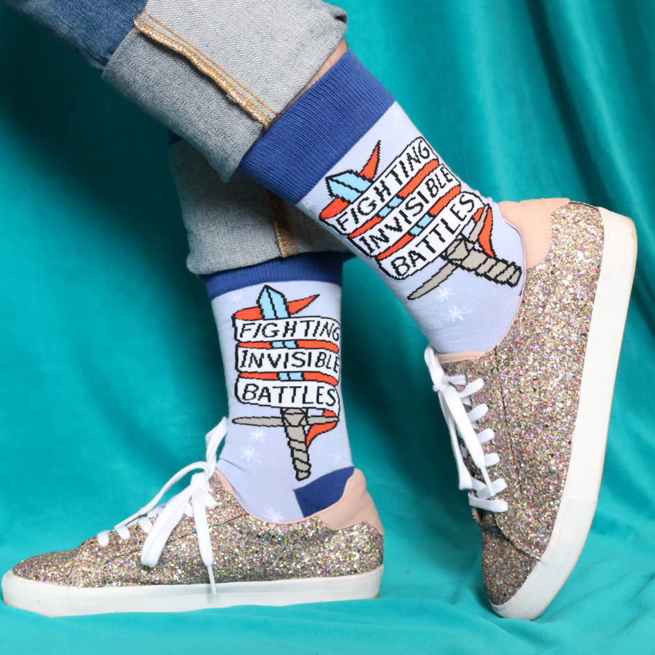 A pair of socks being worn with glitter shoes. The socks are blue and read Fighting Invisible Battles inside a dagger.