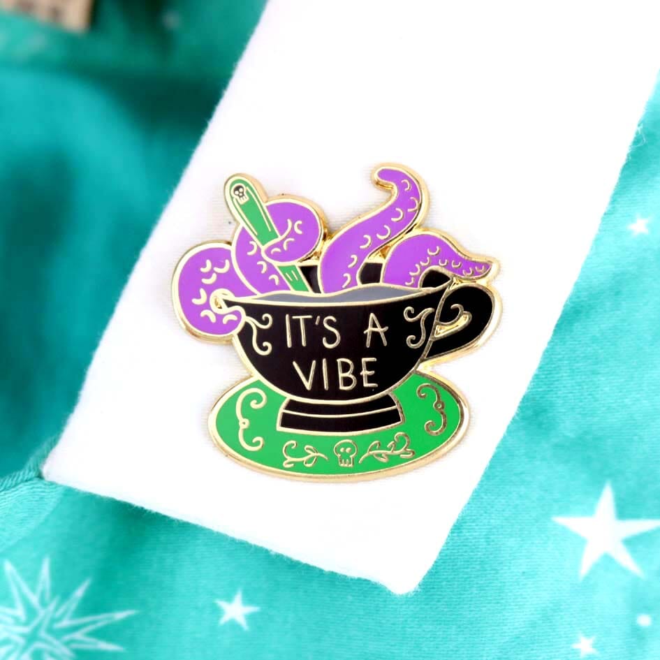 A hard enamel lapel pin being worn on a white collar. The lapel pin is in the shape of a black teacup with purple tenticles coming out. The pin reads It's A Vibe.
