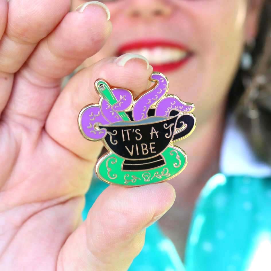 A hard enamel lapel pin being held in a hand. The lapel pin is in the shape of a black teacup with purple tenticles coming out. The pin reads It's A Vibe.