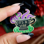 A hard enamel lapel pin being held in a hand. The lapel pin is in the shape of a black teacup with purple tenticles coming out. The pin reads It's A Vibe.