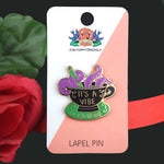 A hard enamel lapel pin displayed on a Jubly-Umph backing card. The lapel pin is in the shape of a black teacup with purple tenticles coming out. The pin reads It's A Vibe.