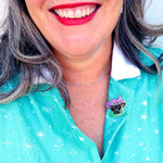 A hard enamel lapel pin being worn by a model with a white collar. The lapel pin is in the shape of a black teacup with purple tenticles coming out. The pin reads It's A Vibe.
