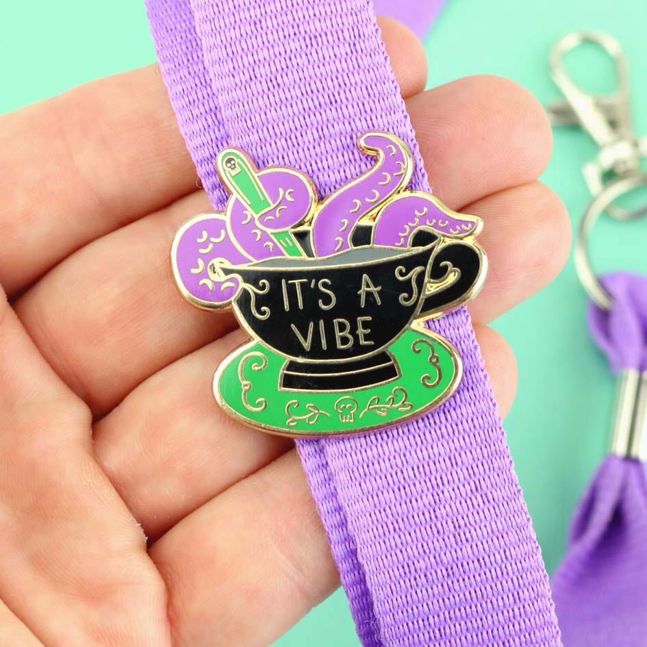 A hard enamel lapel pin displayed on a purple lanyard. The lapel pin is in the shape of a black teacup with purple tenticles coming out. The pin reads It's A Vibe.