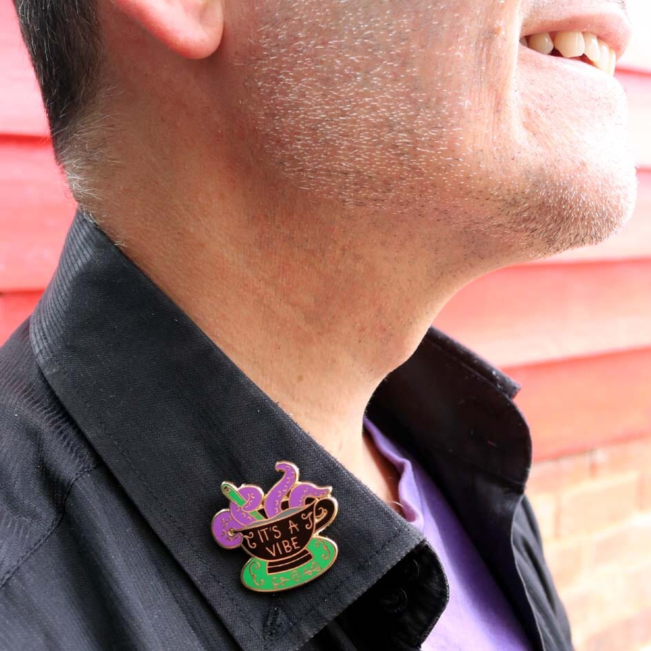 A hard enamel lapel pin being modelled on a black shirt. The lapel pin is in the shape of a black teacup with purple tenticles coming out. The pin reads It's A Vibe.