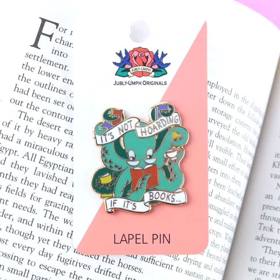 A hard enamel lapel pin displayed on a Jubly-Umph backing card. The pin is in the shape of an Octopus carrying books. The pin reads It's Not Hoarding If It's Books.