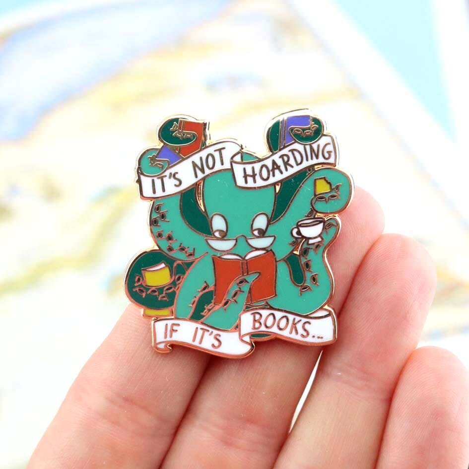 A hard enamel lapel pin being held in a hand. The pin is in the shape of an Octopus carrying books. The pin reads It's Not Hoarding If It's Books.