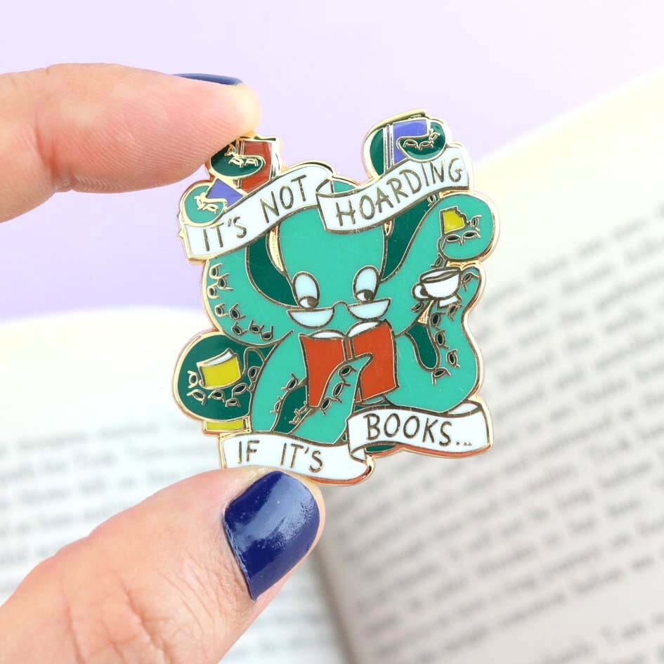 A hard enamel lapel pin being held in a hand. The pin is in the shape of an Octopus carrying books. The pin reads It's Not Hoarding If It's Books.