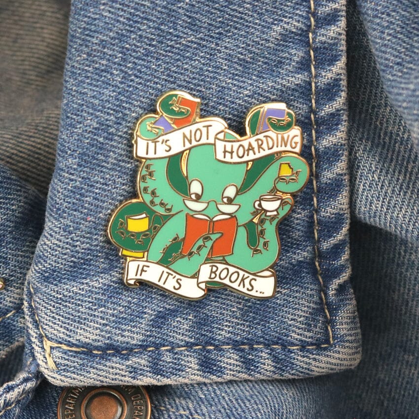 A hard enamel lapel pin being displayed on a denim jacket. The pin is in the shape of an Octopus carrying books. The pin reads It's Not Hoarding If It's Books.
