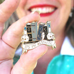 A hard enamel lapel pin being held in a hand. The lapel pin is in the shape of books on a shelf with skulls and candles. The pin reads It's My Aesthetic.