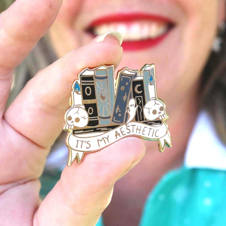A hard enamel lapel pin being held in a hand. The lapel pin is in the shape of books on a shelf with skulls and candles. The pin reads It's My Aesthetic.