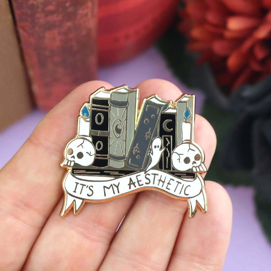 A hard enamel lapel pin being held in a hand. The lapel pin is in the shape of books on a shelf with skulls and candles. The pin reads It's My Aesthetic.