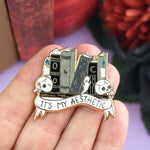A hard enamel lapel pin being held in a hand. The lapel pin is in the shape of books on a shelf with skulls and candles. The pin reads It's My Aesthetic.
