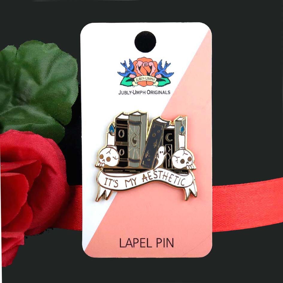 A hard enamel lapel pin on Jubly-Umph card stock. The lapel pin is in the shape of books on a shelf with skulls and candles. The pin reads It's My Aesthetic.