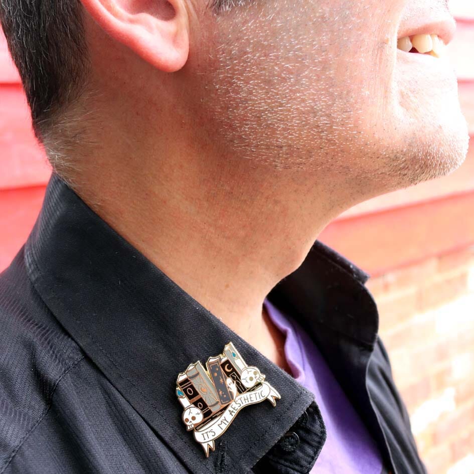 A hard enamel lapel pin being modelled on a black shirt. The lapel pin is in the shape of books on a shelf with skulls and candles. The pin reads It's My Aesthetic.