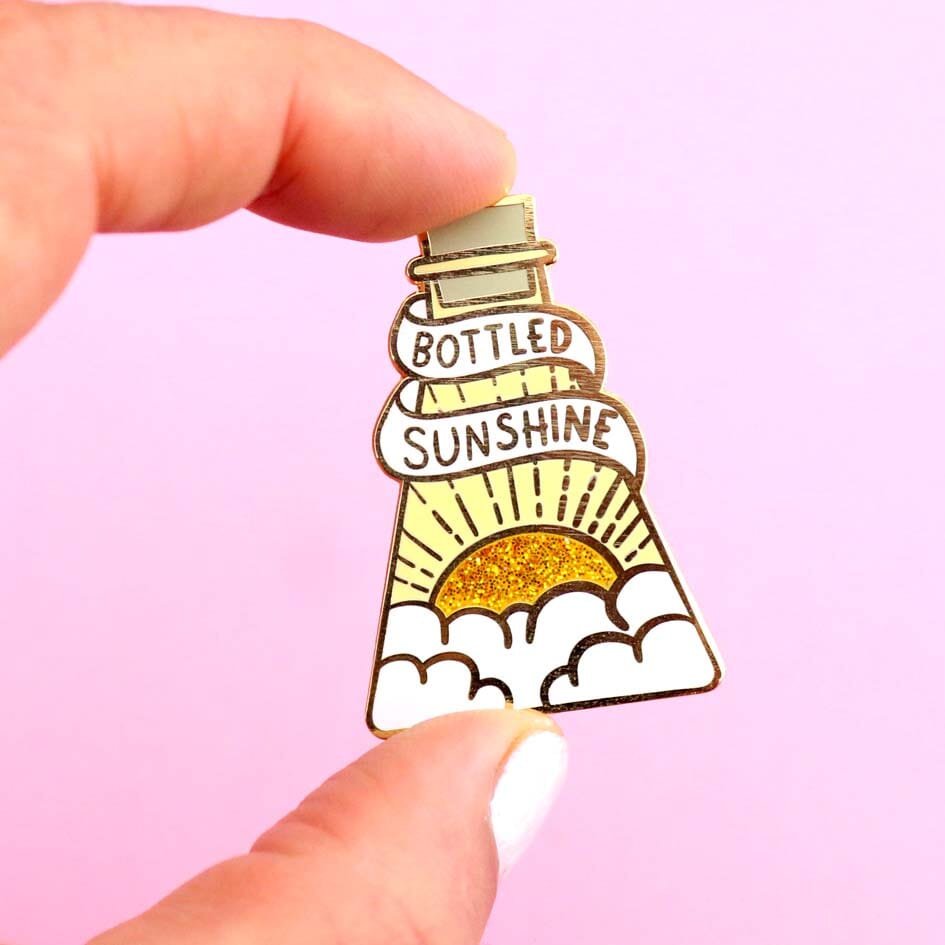 A hard enamel lapel pin being held in a hand. The pin is in the profile of a beaker with a yellow glitter sun. The pin says Bottled Sunshine.