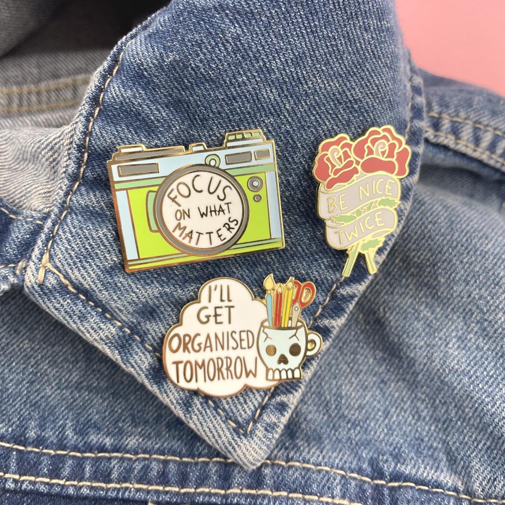 A hard enamel pin on a denim jacket with two other pins. The pin is in the shape of a camera and reads Focus On What Matters.
