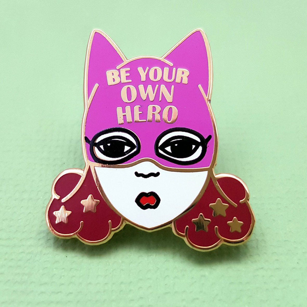 An enamel pin with a picture of a white woman with brown hair and a purple superhero hat. it says "Be  your own hero" on the hat