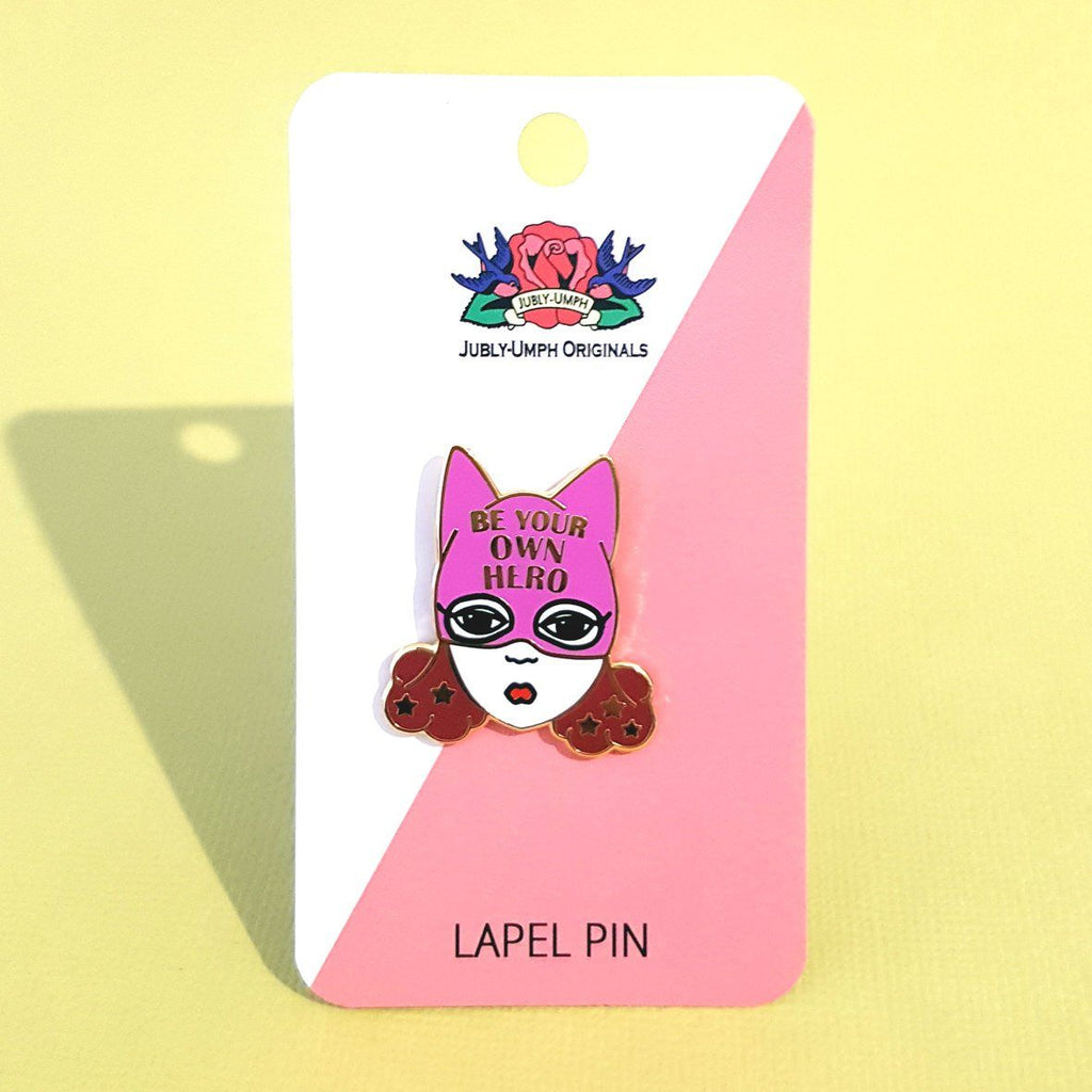 An enamel pin with a picture of a white woman with brown hair and a purple superhero hat. it says "Be  your own hero" on the hat. the pin is shown on a Jubly-Umph backing card against a yellow background