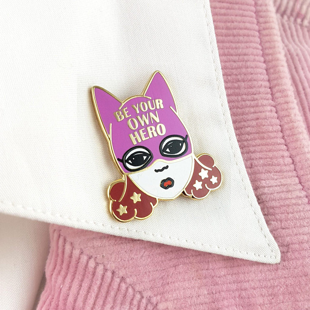 An enamel pin is attached to a white skirt collar. the pin is of a white woman with brown hair and a superhero hat. it says "Be  your own hero" on the hat