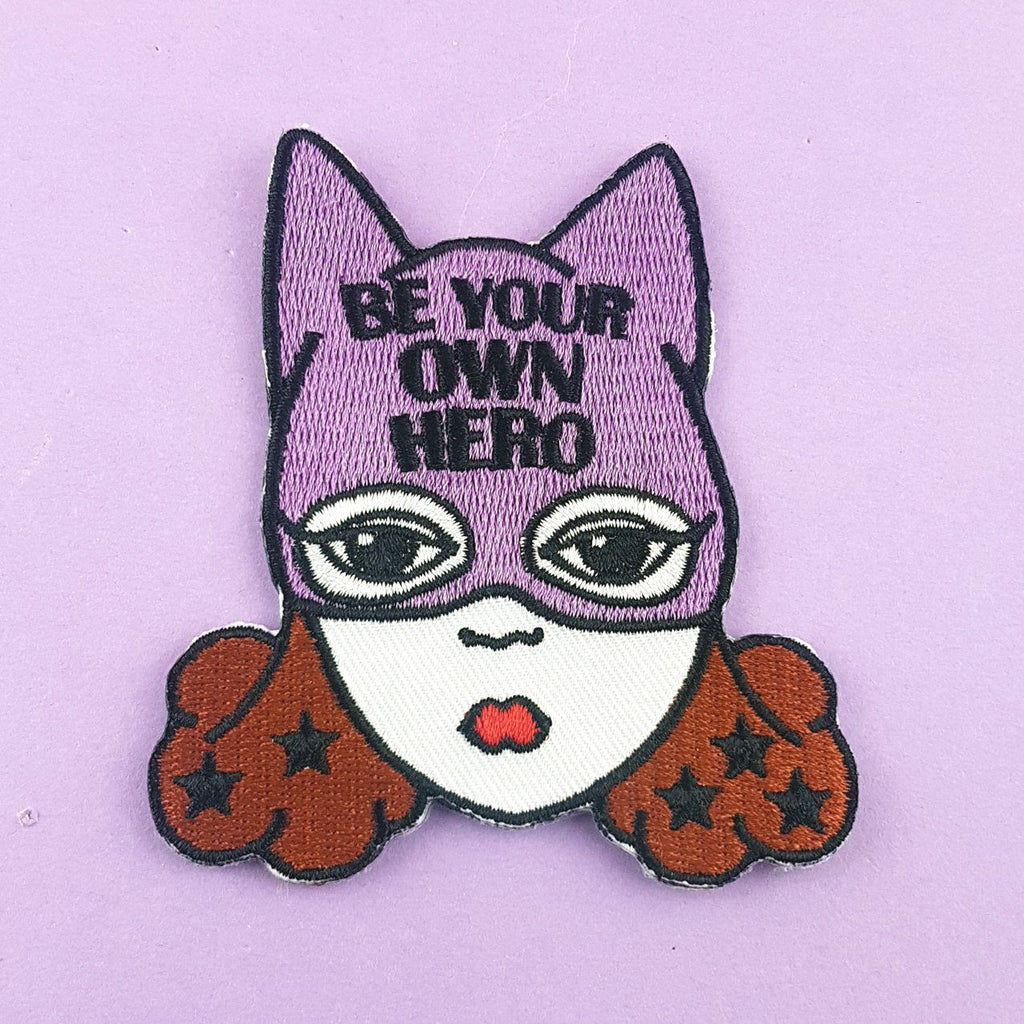 an embroidered iron on patch. The patch is of a white woman with brown hair and a superhero hat. it says "Be  your own hero" on the hat