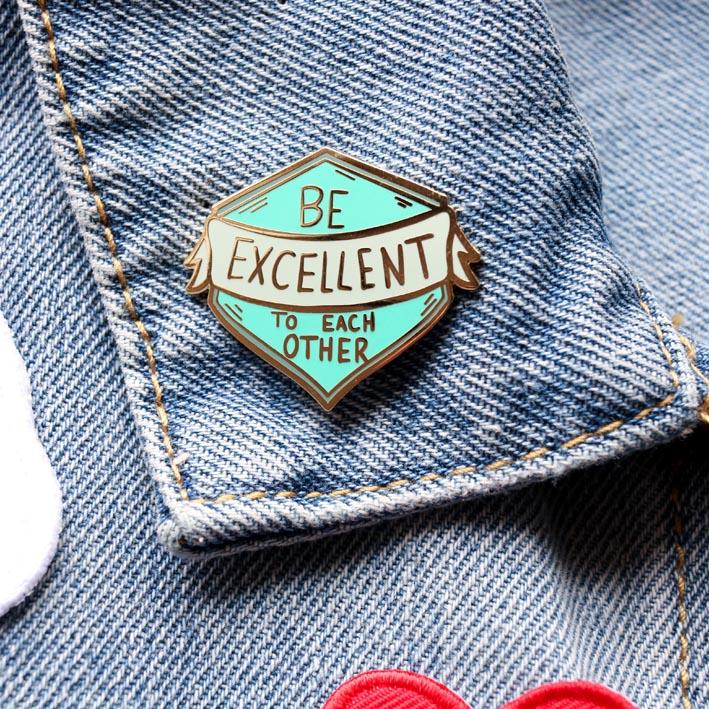 Deninm jacket collar with an enamel pin attached. The pin  is green coloured, in the shape of a shield and reads "Be excellent to each other"