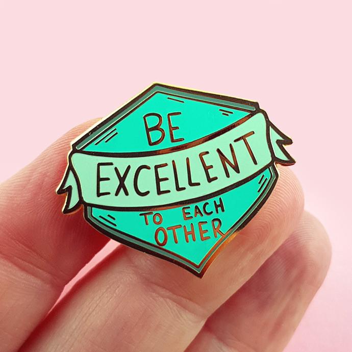 hand holding an Enamel pin in a shield shape with a banner draped on it. The text reads "Be Excellent to each other". The pin colour is mint and sea green