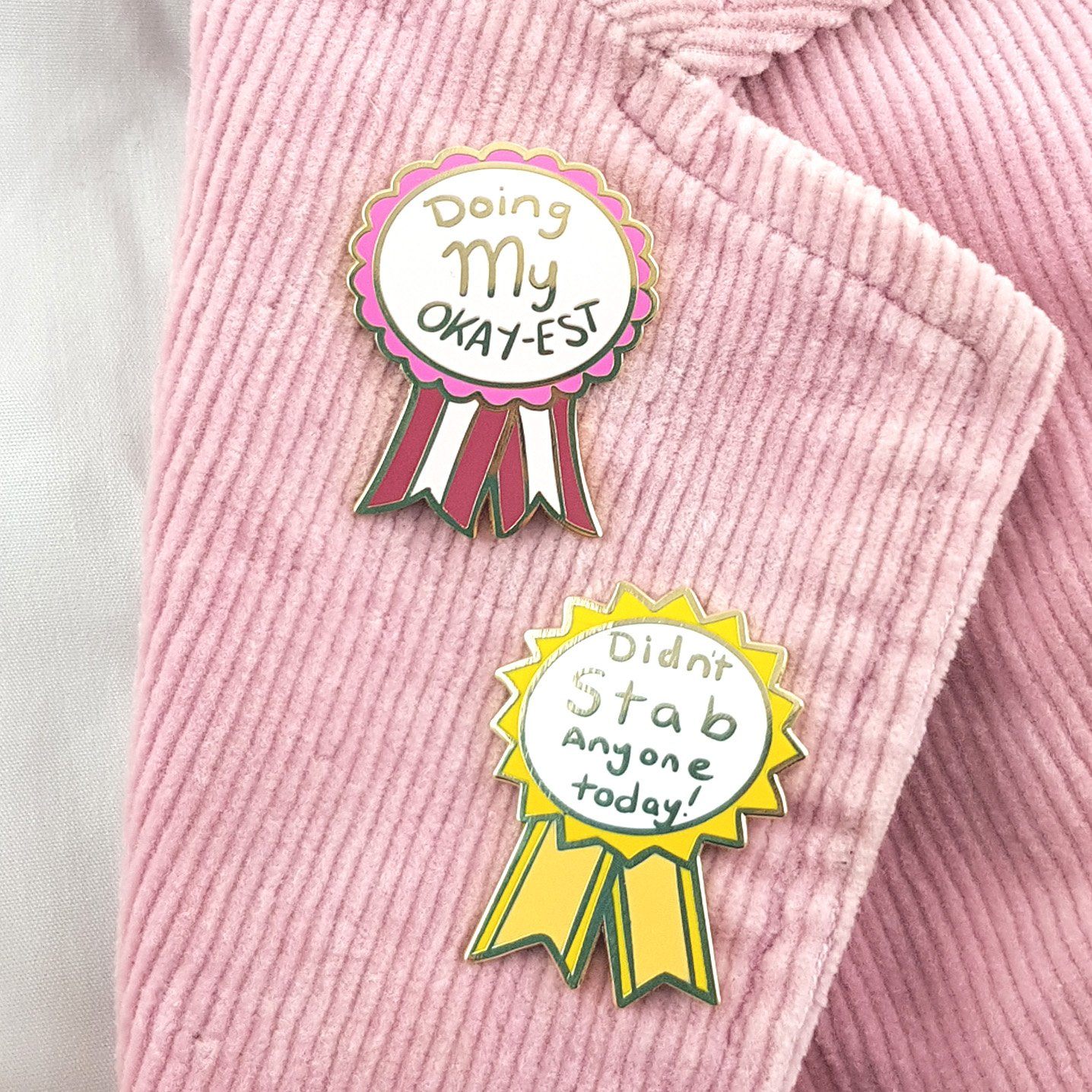 Two hard enamel lapel pins on a pink jacket. The first pin is in the shape of an award ribbon. The ribbon is dark pink and light pink and reads Doing my Okay-est. The second pin is in the shape of an award ribbon and is yellow and white and reads Didn’t Stab Anyone Today!