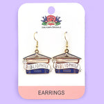 A pair of dangle earrings displayed on Jubly-Umph cardstock. The earrings say Bibliophile in the middle of a stack of blue books.
