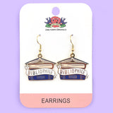 A pair of dangle earrings displayed on Jubly-Umph cardstock. The earrings say Bibliophile in the middle of a stack of blue books.