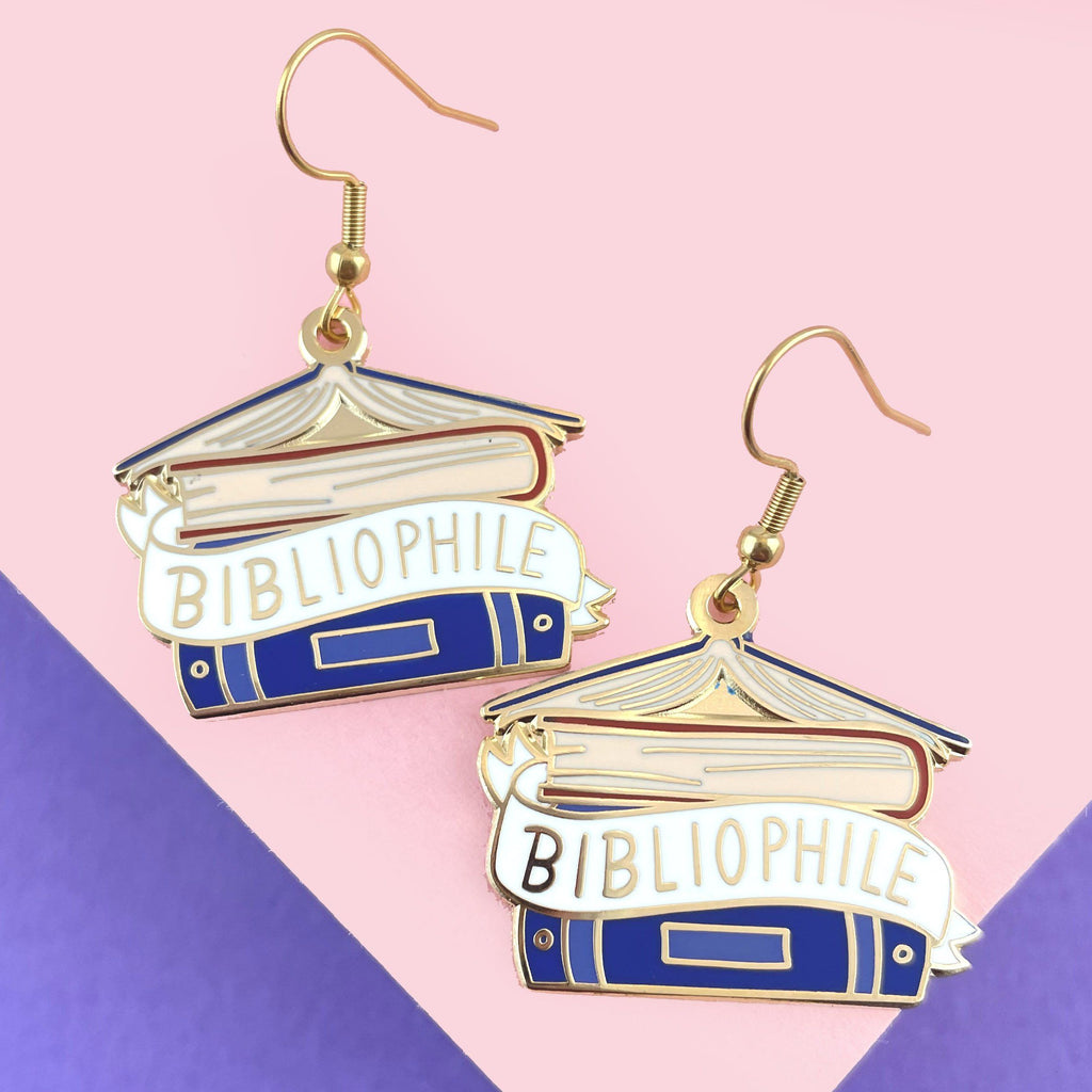 A pair of dangle earrings displayed on a pink background. The earrings say Bibliophile in the middle of a stack of blue books.