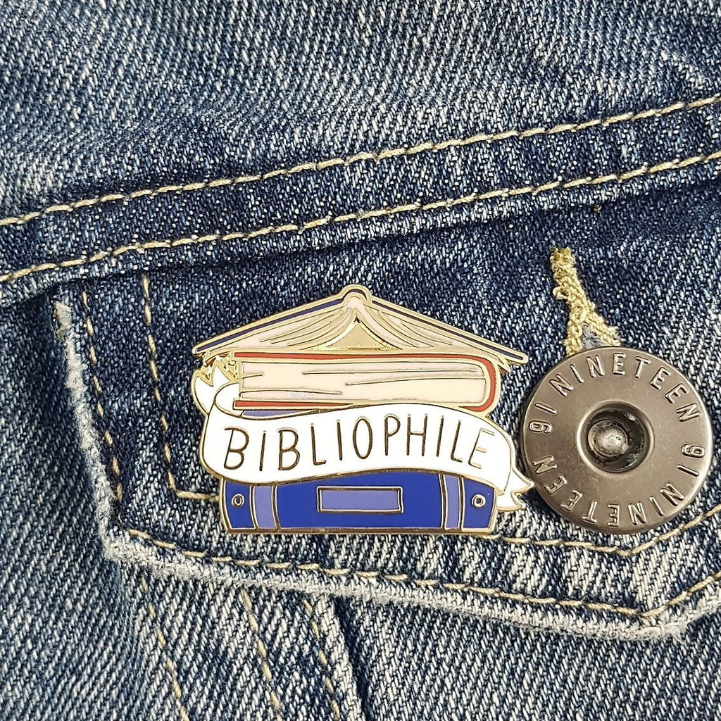 Hard enamel lapel pin depicting a stack of blue and red book spines, with a white banner wrapped around displaying the word 'bibliophile' pinned to the pocket of a denim jacket.