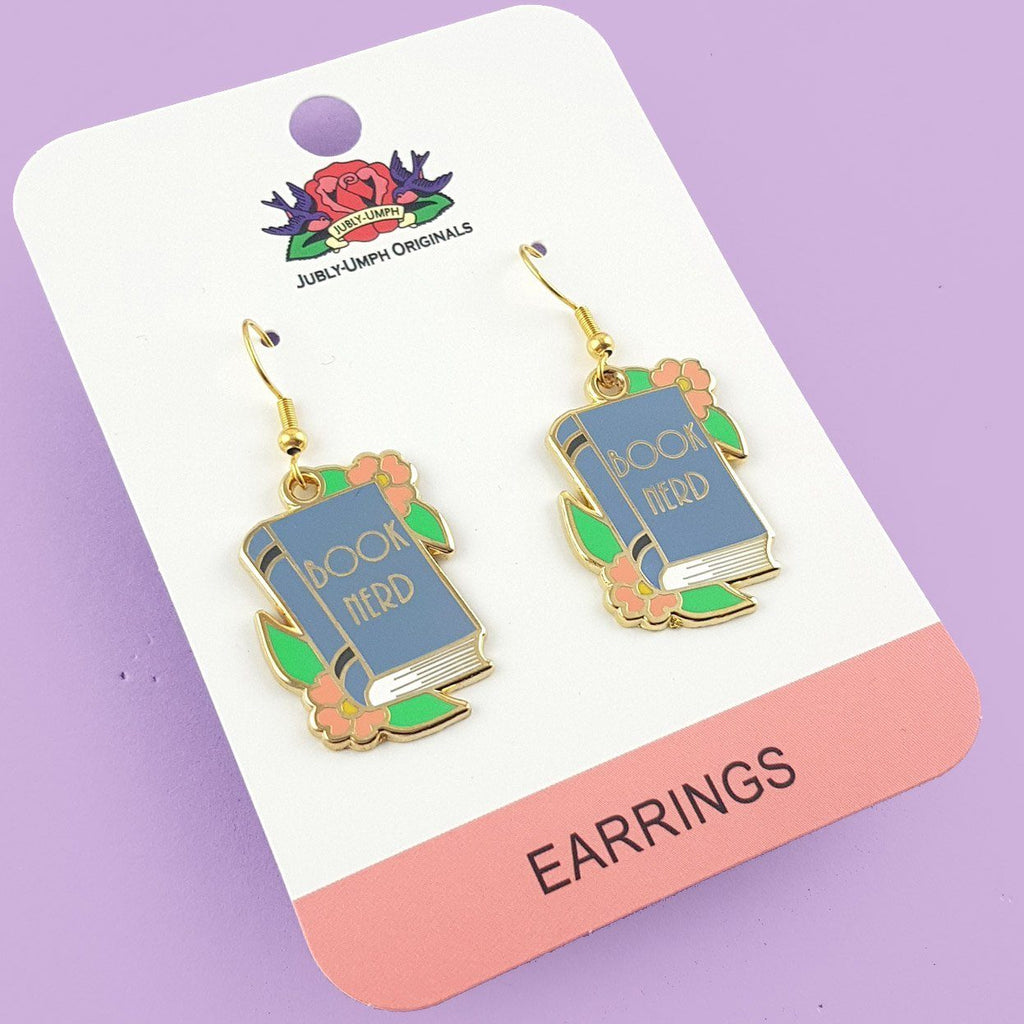 A pair of dangle earrings displayed on on Jubly-Umph cardstock. The earrings say Book Nerd in the middle of a blue book.